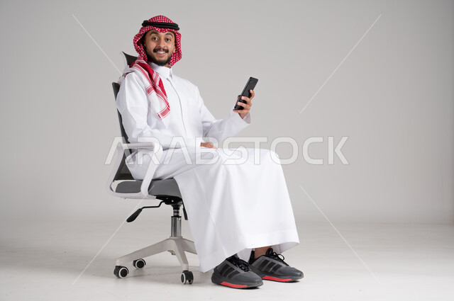 استخدام الأجهزة والتقنيات الحديثة، بورتريه لرجل عربي خليجي سعودي يرتدي الشماغ والثوب التقليدي يجلس على كرسي دوار يمسك الجوال وينظر إلى الكاميرا بإيماءات السرور، مفهوم الأناقة والرجولة، الاهتمام بالمظهر الخارجي والثقة بالنفس، الجسم بالطول الكامل، خلفية رمادية