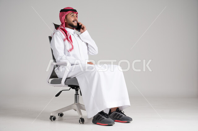 إجراء مكالمة هاتفية باستخدام الهاتف المحمول، الاستفادة من الأجهزة والتقنيات الحديثة، بورتريه لرجل عربي خليجي سعودي يرتدي الشماغ والثوب التقليدي يجلس على كرسي دوار يمسك الجوال بيده، الاطمئنان على الأهل والأصدقاء وسماع أخبارهم، الجسم بالطول الكامل، خلفية رمادية