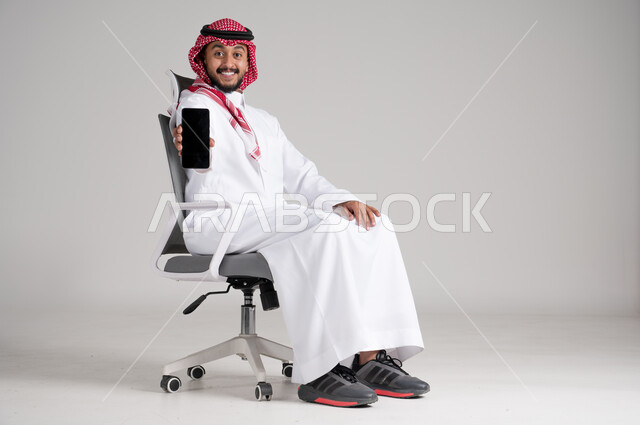 Holding mobile phone and showing blank black screen, portrait of a Saudi Arabian Gulf man wearing traditional keffiyeh and thobe sitting on a swivel chair and looking at the camera with gestures of pleasure, concept of elegance and masculinity, concern for appearance and self-confidence, full body, gray background