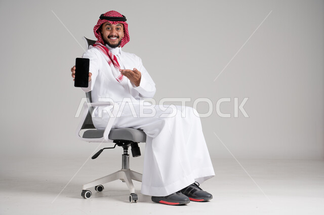الإمساك بالهاتف المحمول وعرض شاشة سوداء فارغة، بورتريه لرجل عربي خليجي سعودي يرتدي الشماغ والثوب التقليدي يجلس على كرسي دوار ويشير إلى الجوال بإيماءات السرور، مفهوم الأناقة والرجولة، الاهتمام بالمظهر الخارجي والثقة بالنفس، الجسم بالطول الكامل، خلفية رمادية