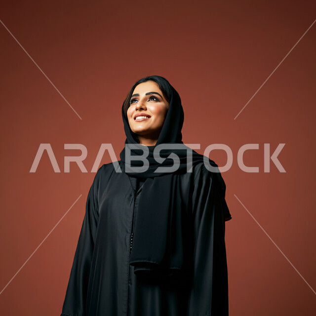  تعابير البهجة والسرور والسعادة، الاهتمام بالأناقة والمظهر الخارجي، مفهوم الأنوثة والنعومة، بورتريه مقرب لامرأة عربية خليجية سعودية محجبة مبتسمة ترتدي العباءة السوداء تنظر إلى الأعلى  بإيماءات الثقة بالنفس والفرح، خلفية حمراء