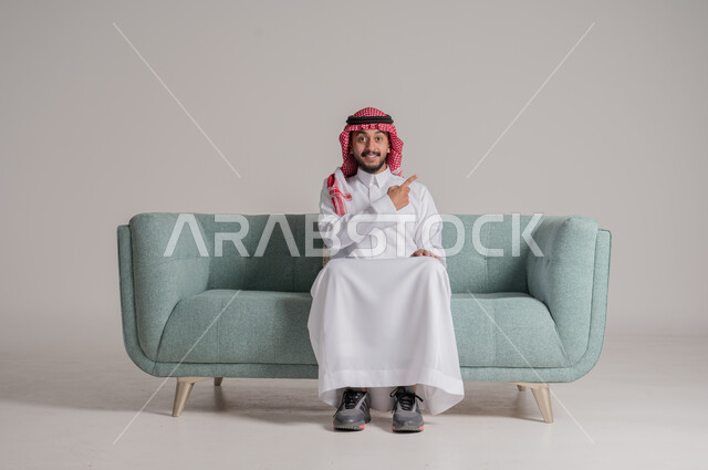 تحديد إلى شيء ما بإيماءات السرور, بورتريه  لرجل عربي خليجي سعودي مبتسم يرتدي الثوب التقليدي والشماغ يجلس على كنبة مريحة، الإشارة بأصابع السبابة إلى هدف محدد، النظر إلى الكاميرا بتعابير السعادة، خلفية رمادية