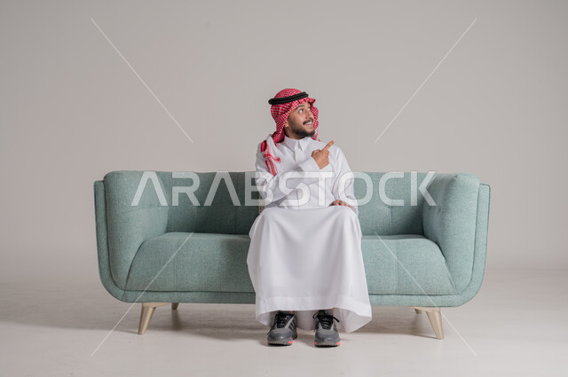 تعابير التأكد والتحديد, تحديد شيء ما بإيماءات السرور ,بورتريه  لرجل عربي خليجي سعودي مبتسم يرتدي الثوب التقليدي والشماغ يجلس على كنبة مريحة، الإشارة بأصابع السبابة إلى هدف محدد، خلفية رمادية