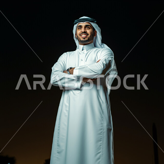 The concept of handsomeness and masculinity, elegance and interest in external appearance, close-up portrait of a smiling Saudi Arabian Gulf man wearing a white ghutra and traditional thobe standing with crossed hands expressions, standing straight and looking at the camera with gestures of pleasure and self-confidence, black background