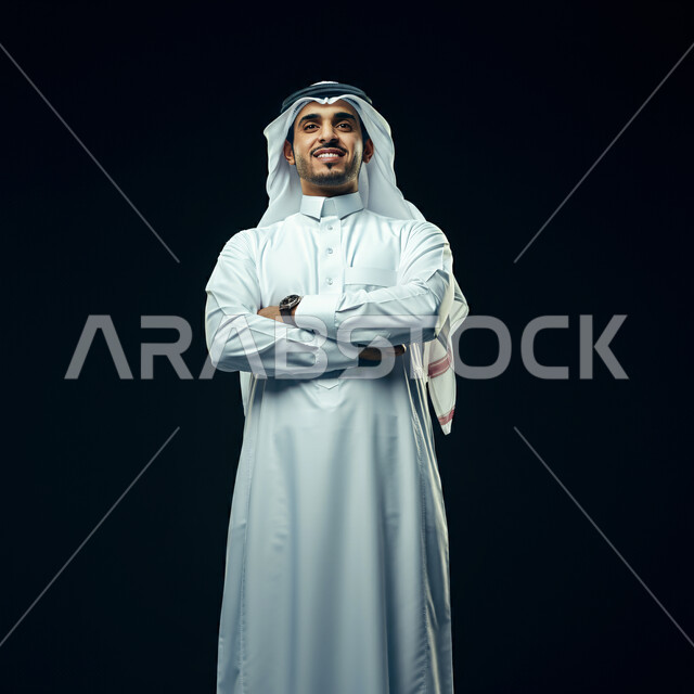 مفهوم الوسامة والرجولة، الأناقة والاهتمام بالمظهر الخارجي، بورتريه مقرب لرجل عربي خليجي سعودي مبتسم يرتدي الغترة البيضاء والثوب التقليدي يقف بتعابير أيدي متقاطعة، الوقوف بإستقامة والنظر الى الكاميرا بايماءات السرور والثقة بالنفس، خلفية سوداء
