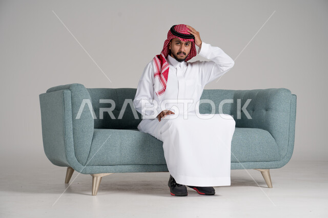 آلام الصداع النصفي والشقيقة، التعرض لنوبات وجع الرأس، بورتريه لرجل عربي خليجي سعودي يرتدي الشماغ والثوب التقليدي يجلس على كنبة مريحة في غرفة المعيشة ويضع يده على رأسه بإيماءات التعب وعدم الراحة، الجسم بالطول الكامل، خلفية رمادية