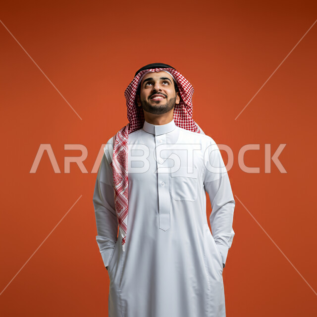 الوقوف بإستقامة والنظر الى الاعلى بايماءات السرور والثقة بالنفس، مفهوم الرجولة والأناقة والاهتمام بالمظهر الخارجي، بورتريه مقرب لرجل سعودي عربي خليجي مبتسم يرتدي الثوب التقليدي والشماغ يضع يديه في جيبه، خلفية برتقالية
