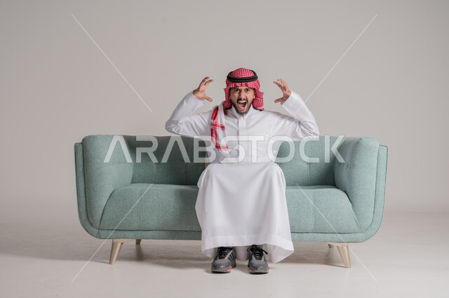 رفع اليدين في الهواء بتعابير الغضب، النظر للكاميرا بإيماءات الانزعاج وعدم الرضا، بورتريه لرجل عربي خليجي سعودي يرتدي الثوب التقليدي والشماغ يجلس على كنبة مريحة يرفع كفيه بتذمر وسخط، الجسم بالطول الكامل، خلفية رمادية