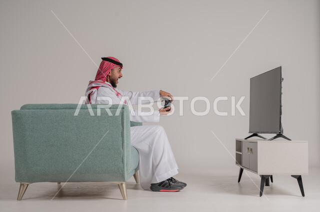 استخدام التقنيات وأجهزة التكنولوجيا الحديثة، الاستمتاع بألعاب الفيديو في وقت الفراغ، بورتريه من الجانب لرجل عربي خليجي سعودي يرتدي الشماغ والثوب التقليدي يجلس على كنبة مريحة في غرفة المعيشة يمسك عصا التحكم بإيماءات الاندماج، الجسم بالطول الكامل، خلفية رمادية