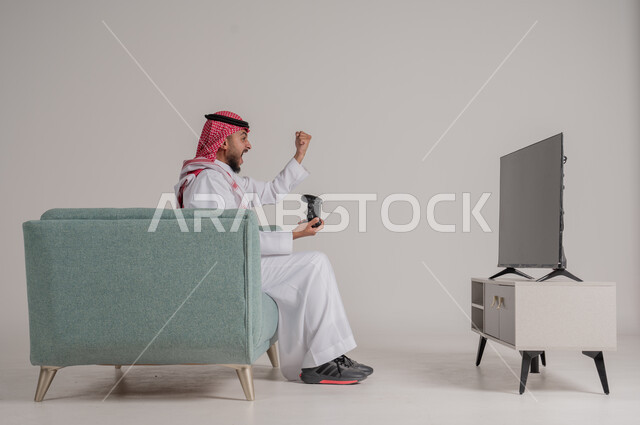 استخدام التقنيات وأجهزة التكنولوجيا الحديثة، الاستمتاع بألعاب الفيديو في وقت الفراغ، بورتريه من الجانب لرجل عربي خليجي سعودي يرتدي الشماغ والثوب التقليدي يجلس على كنبة مريحة في غرفة المعيشة يمسك عصا التحكم بإيماءات الاندماج، الجسم بالطول الكامل، خلفية رمادية