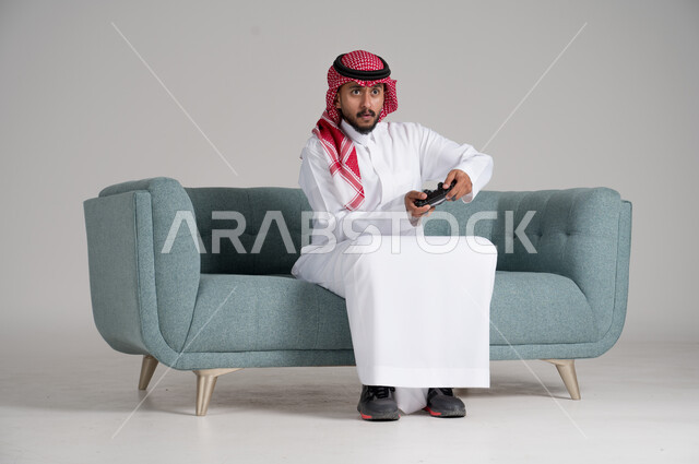استخدام التقنيات والأجهزة التكنولوجية الحديثة، الاستمتاع بألعاب الفيديو في وقت الفراغ، بورتريه لرجل عربي خليجي سعودي يرتدي الشماغ والثوب التقليدي يجلس على كنبة مريحة في غرفة المعيشة يمسك عصا التحكم بإيماءات الاندماج والتركيز، الجسم بالطول الكامل، خلفية رمادية