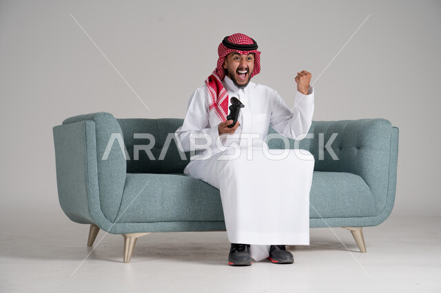 الاستمتاع بألعاب الفيديو في أوقات الفراغ، استخدام التقنيات والأجهزة التكنولوجية الحديثة، بورتريه لرجل عربي خليجي سعودي يرتدي الشماغ والثوب التقليدي يجلس على كنبة مريحة في غرفة المعيشة يمسك عصا التحكم ويرفع يده بحماس واندماج، الجسم بالطول الكامل، خلفية رمادية
