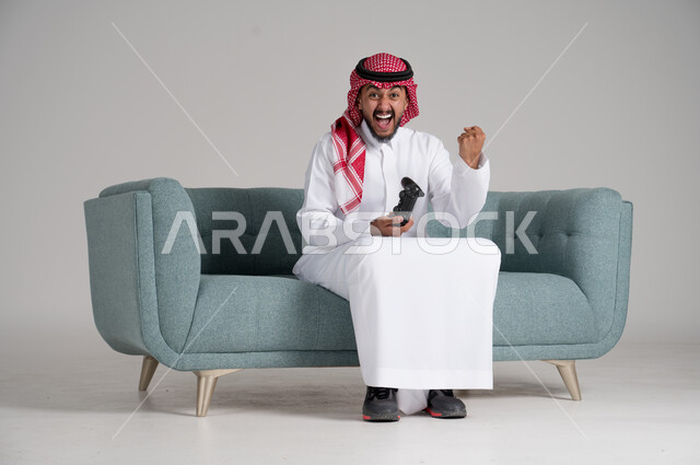 الاستمتاع بألعاب الفيديو في أوقات الفراغ، استخدام التقنيات والأجهزة التكنولوجية الحديثة، بورتريه لرجل عربي خليجي سعودي يرتدي الشماغ والثوب التقليدي يجلس على كنبة مريحة في غرفة المعيشة يمسك عصا التحكم ويرفع يده في الهواء بحماس، الجسم بالطول الكامل، خلفية رمادية
