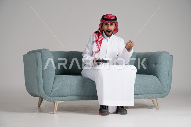 الاستمتاع بألعاب الفيديو في أوقات الفراغ، استخدام التقنيات والأجهزة التكنولوجية الحديثة، بورتريه لرجل عربي خليجي سعودي يرتدي الشماغ والثوب التقليدي يجلس على كنبة مريحة في غرفة المعيشة يمسك عصا التحكم ويرفع يده في الهواء بحماس، الجسم بالطول الكامل، خلفية رمادية