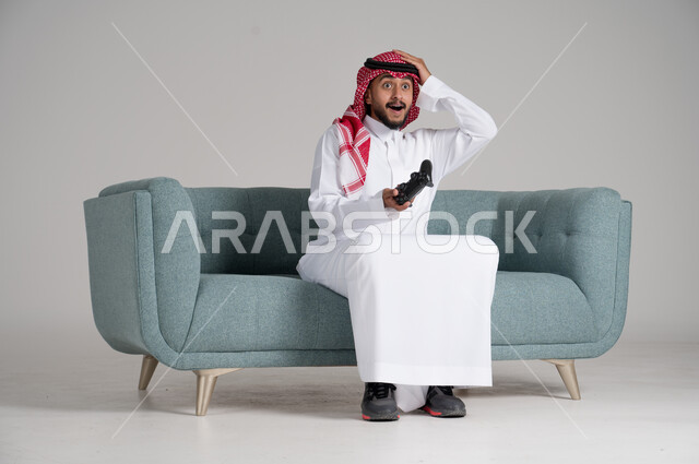 الاستمتاع بألعاب الفيديو في أوقات الفراغ، استخدام التقنيات والأجهزة التكنولوجية الحديثة، بورتريه لرجل عربي خليجي سعودي يرتدي الشماغ والثوب التقليدي يجلس على كنبة مريحة في غرفة المعيشة يمسك عصا التحكم ويضع يده على رأسه، الجسم بالطول الكامل، خلفية رمادية