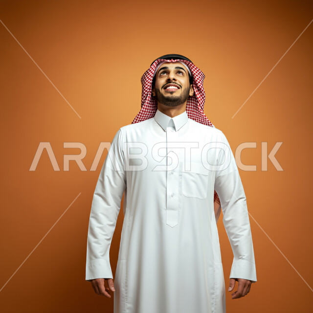 مفهوم الرجولة والأناقة والاهتمام بالمظهر الخارجي، بورتريه مقرب لرجل سعودي عربي خليجي مبتسم يرتدي الثوب التقليدي والشماغ يضع يديه على جانبه، الوقوف بإستقامة والنظر الى الاعلى بايماءات السرور و الانضباط والثقة بالنفس، خلفية بنية