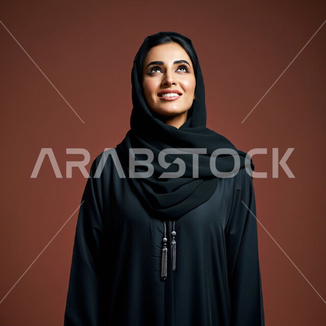 مفهوم الأنوثة والنعومة، بورتريه مقرب لامرأة عربية خليجية سعودية محجبة مبتسمة ترتدي العباءة السوداء تنظر إلى الأعلى  بإيماءات الثقة بالنفس والفرح، الاهتمام بالأناقة والمظهر الخارجي، تعابير السرور والسعادة، خلفية بنية