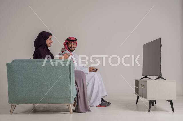 التحدي والاستمتاع بألعاب الفيديو في المنزل بأوقات الفراغ، استخدام أجهزة التكنولوجيا الحديثة، بورتريه من الجانب لزوجين عربيين خليجيين سعوديين يرتديان الملابس التقليدية يجلسان على كنبة مريحة في غرفة المعيشة يمسكان عصي التحكم، الجسم بالطول الكامل، خلفية رمادية