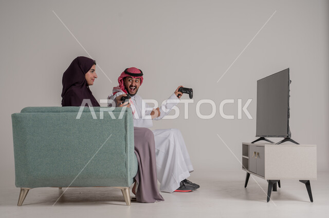 التحدي والاستمتاع بألعاب الفيديو في المنزل بأوقات الفراغ، استخدام أجهزة التكنولوجيا الحديثة، بورتريه من الجانب لزوجين عربيين خليجيين سعوديين يرتديان الملابس التقليدية يجلسان على كنبة مريحة في غرفة المعيشة يمسكان عصي التحكم، الجسم بالطول الكامل، خلفية رمادية