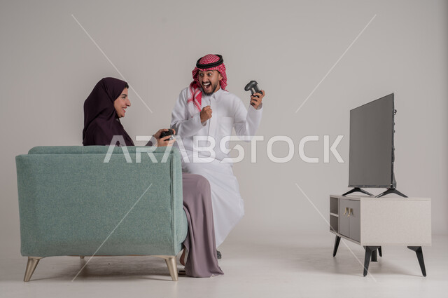 التحدي والاستمتاع بألعاب الفيديو في المنزل بأوقات الفراغ، استخدام أجهزة التكنولوجيا الحديثة، بورتريه من الجانب لزوجين عربيين خليجيين سعوديين يرتديان الملابس التقليدية يجلسان على كنبة مريحة في غرفة المعيشة يمسكان عصي التحكم، الجسم بالطول الكامل، خلفية رمادية