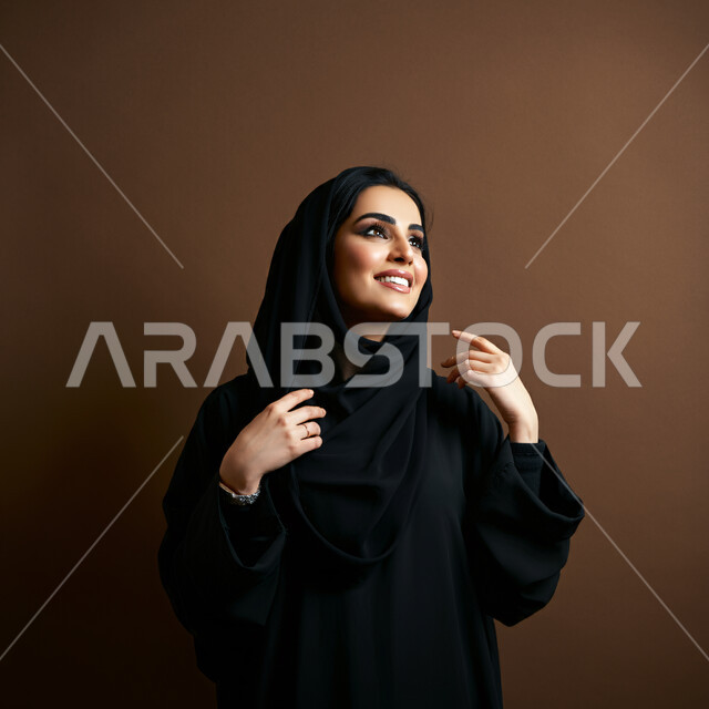 Expressions of joy, pleasure and happiness, interest in elegance and appearance, concept of femininity and softness, close-up portrait of a smiling veiled Saudi Arabian Gulf woman wearing a black abaya looking up with gestures of self-confidence and joy, brown background