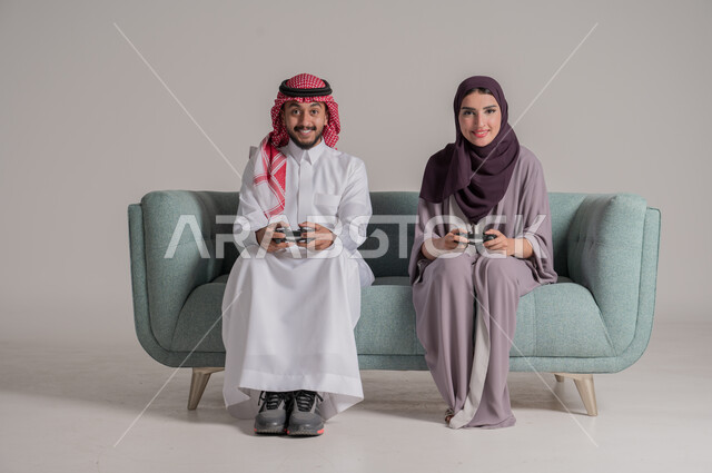 اللعب بألعاب الفيديو في المنزل برفقة الزوجة، استخدام أجهزة التكنولوجيا الحديثة، بورتريه لزوجين عربيين خليجيين سعوديين يرتديان الملابس التقليدية يجلسان على كنبة مريحة في غرفة المعيشة يمسكان عصي التحكم بإيماءات التحدي والتركيز، الجسم بالطول الكامل، خلفية رمادية