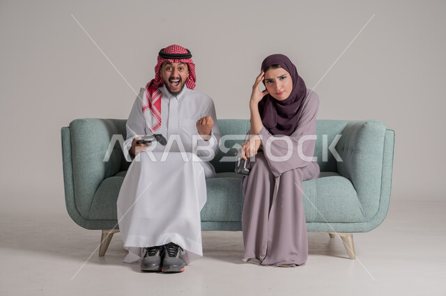 Playing video games at home with wife, using modern technology devices, portrait of a Saudi Arabian Gulf couple wearing traditional clothes sitting on a comfortable sofa in the living room holding joysticks with gestures of challenge and concentration, full body, gray background