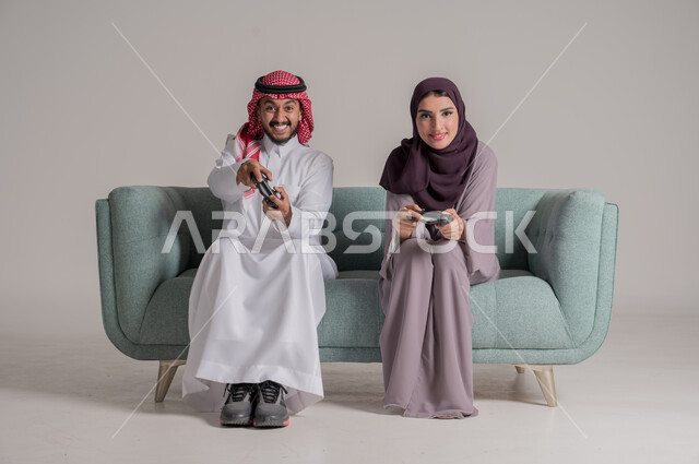 اللعب بألعاب الفيديو في المنزل برفقة الزوجة، استخدام أجهزة التكنولوجيا الحديثة، بورتريه لزوجين عربيين خليجيين سعوديين يرتديان الملابس التقليدية يجلسان على كنبة مريحة في غرفة المعيشة يمسكان عصي التحكم بإيماءات التحدي والتركيز، الجسم بالطول الكامل، خلفية رمادية