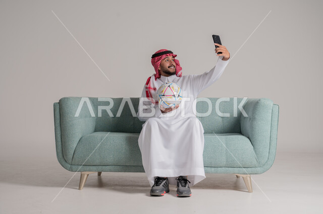 Taking a souvenir selfie, using modern and advanced technical devices, competition, challenge and encouragement, portrait of a Saudi Arabian Gulf man wearing a traditional thobe and shemagh sitting on a comfortable sofa holding a football and a mobile phone, gray background