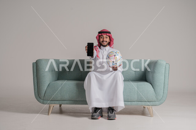 المنافسة والتحدي والتشجيع, بورتريه لرجل عربي سعودي خليجي يرتدي الثوب التقليدي والشماغ يجلس على كنبة مريحة يمسك بيده كرة القدم والهاتف المحمول وينظر الى الكاميرا، عرض شاشة سوداء فارغة على الموبايل، استخدام الأجهزة التقنية الحديثة والتطورة، خلفية رمادية
