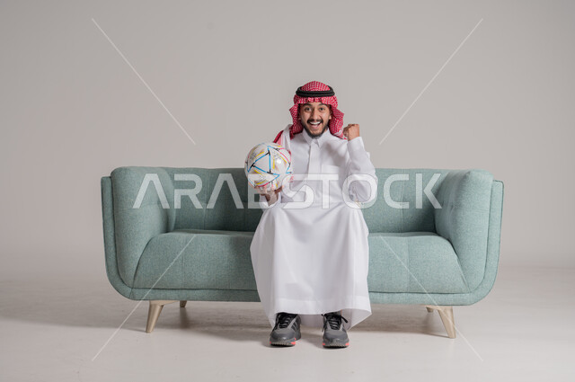 Interaction and integration with the match, looking at the camera with expressions of pleasure, joy at winning and triumph, portrait of a Saudi Arabian Gulf man wearing traditional thobe and shemagh sitting on a sofa holding the ball and raising his hand with his palm closed cheering, facial and hand gestures indicating happiness, gray background
