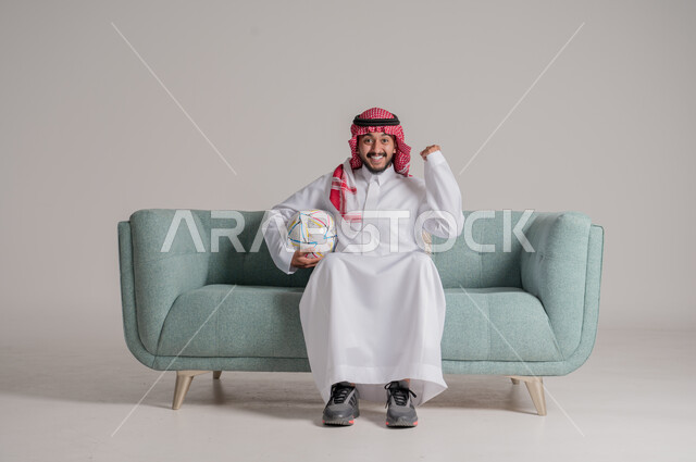 إيماءات وجه ويدين تدل على السعاده, التفاعل والإندماج مع المباراة, النظر الى الكاميرا بتعابير السرور، الفرح بالفوز والإنتصار, بورتريه لرجل عربي سعودي خليجي يرتدي الثوب التقليدي والشماغ يجلس على كنبة يحمل الكرة ويرفع يده مضموم الكف يقوم بالتشجيع, خلفية رمادية