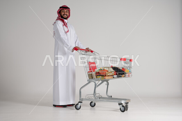 الاستمتاع بالتسوق في المولات والمراكز التجارية، شراء المستلزمات والحاجيات الضرورية، الاستفادة من الخصومات على المنتجات، بورتريه لرجل عربي خليجي سعودي يرتدي الشماغ والثوب التقليدي ويجر عربة التسوق أمامه بإيماءات السعادة، الجسم بالطول الكامل، خلفية رمادية