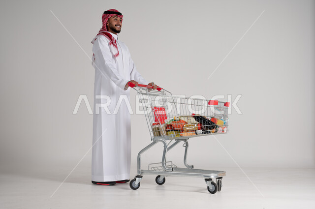 الاستمتاع بالتسوق في المولات والمراكز التجارية، شراء المستلزمات والحاجيات الضرورية، الاستفادة من الخصومات على المنتجات، بورتريه لرجل عربي خليجي سعودي يرتدي الشماغ والثوب التقليدي ويجر عربة التسوق أمامه بإيماءات السعادة، الجسم بالطول الكامل، خلفية رمادية