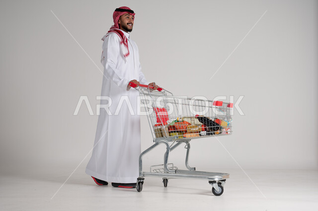الاستمتاع بالتسوق في المولات والمراكز التجارية، شراء المستلزمات والحاجيات الضرورية، الاستفادة من الخصومات على المنتجات، بورتريه لرجل عربي خليجي سعودي يرتدي الشماغ والثوب التقليدي ويجر عربة التسوق أمامه بإيماءات السعادة، الجسم بالطول الكامل، خلفية رمادية