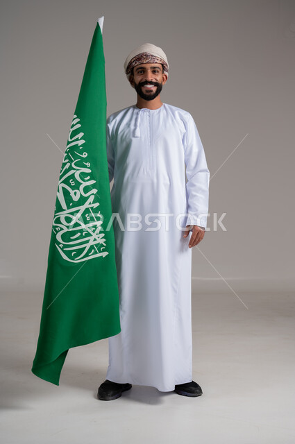 ذكرى تأسيس الدولة السعودية الأولى 22 فبراير، الفخر والاعتزاز بإنجازات الوطن،  شعار الهوية الوطنية وراية الإسلام والتوحيد، بورتريه لرجل عربي خليجي عماني مبتسم يرتدي الدشداشة والعمامة يحمل بيده علم التأسيس, الطول الكامل للجسم، خلفية رمادية