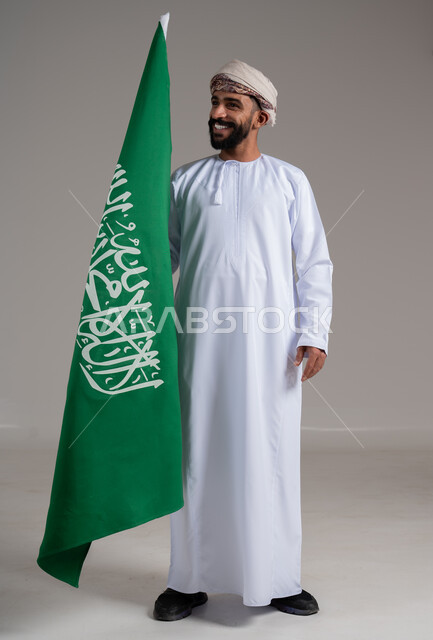 الفخر والاعتزاز بإنجازات الوطن، شعار الهوية الوطنية وراية الإسلام والتوحيد، بورتريه لرجل عربي خليجي عماني مبتسم يرتدي الدشداشة والعمامة يحمل بيده علم التأسيس, ذكرى تأسيس الدولة السعودية الأولى 22 فبراير، الطول الكامل للجسم، خلفية رمادية
