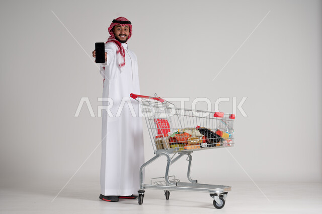 الوقوف وعرض شاشة جوال سوداء فارغة، الاستمتاع بالتسوق في المراكز التجارية، الاستفادة من فترة التنزيلات والحسومات، بورتريه لرجل عربي خليجي سعودي يرتدي الشماغ والثوب التقليدي يمسك الهاتف المحمول بيده وأمامه عربة المشتريات، الجسم بالطول الكامل، خلفية رمادية