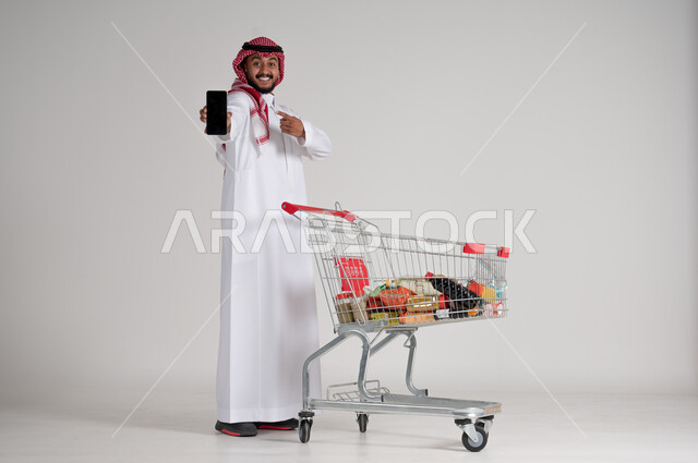 الوقوف والإشارة إلى شاشة الجوال السوداء الفارغة، الاستمتاع بالتسوق في المراكز التجارية، الاستفادة من فترة التنزيلات، بورتريه لرجل عربي خليجي سعودي يرتدي الشماغ والثوب التقليدي يمسك الهاتف المحمول بيده وأمامه عربة المشتريات، الجسم بالطول الكامل، خلفية رمادية