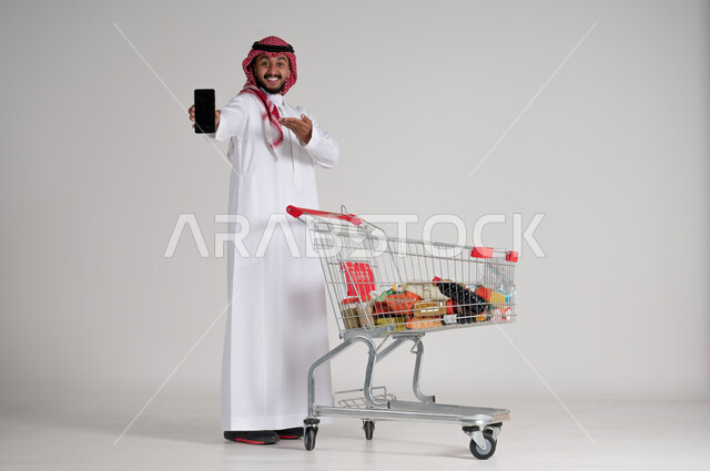 Standing and pointing at blank black mobile screen, enjoying shopping in malls, taking advantage of sales period, portrait of a Saudi Arabian Gulf man wearing traditional shemagh and thobe holding mobile phone in hand and shopping cart in front of him, full body, gray background