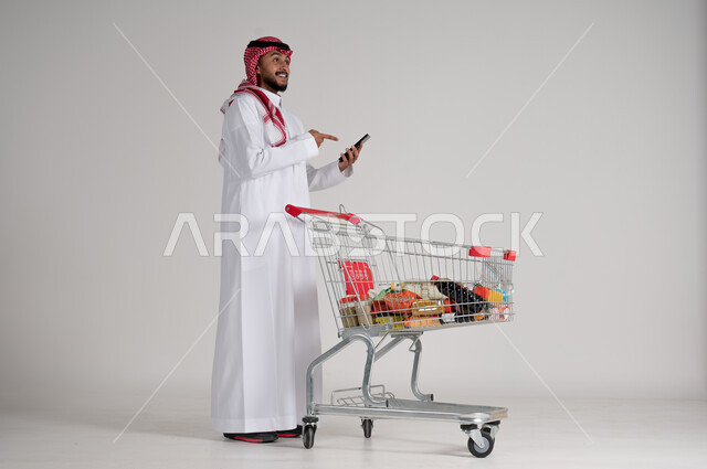 الإمساك بالهاتف المحمول والإشارة إليه، الاستمتاع بالتسوق في المولات والمراكز التجارية، الاستفادة من فترة التنزيلات والحسومات، بورتريه لرجل عربي خليجي سعودي يرتدي الشماغ والثوب التقليدي وأمامه عربة المشتريات والمواد الغذائية، الجسم بالطول الكامل، خلفية رمادية