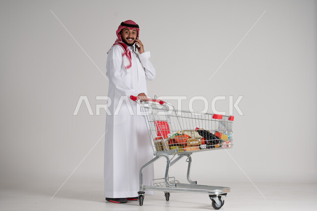 إجراء المكالمات الهاتفية باستخدام الهاتف المحمول، الاستمتاع بالتسوق في المولات والمراكز التجارية، الاستفادة من فترة التنزيلات والحسومات، بورتريه لرجل عربي خليجي سعودي يرتدي الشماغ والثوب التقليدي وأمامه عربة المشتريات، الجسم بالطول الكامل، خلفية رمادية