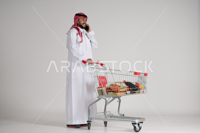 إجراء المكالمات الهاتفية باستخدام الهاتف المحمول، الاستمتاع بالتسوق في المولات والمراكز التجارية، الاستفادة من فترة التنزيلات والحسومات، بورتريه لرجل عربي خليجي سعودي يرتدي الشماغ والثوب التقليدي وأمامه عربة المشتريات، الجسم بالطول الكامل، خلفية رمادية
