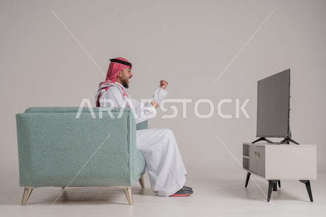 متابعة مباراة كرة القدم بتركيز وتمعن، بورتريه من الجانب لرجل عربي خليجي سعودي يجلس على كنبة مريحة امام التلفاز ويشاهد لعب فريقه المفضل، رفع اليدين بإيماءات الحماس والسعادة لإحراز هدف، تنمية شغف الاهتمامات الرياضية، الجسم بالطول الكامل، خلفية رمادية