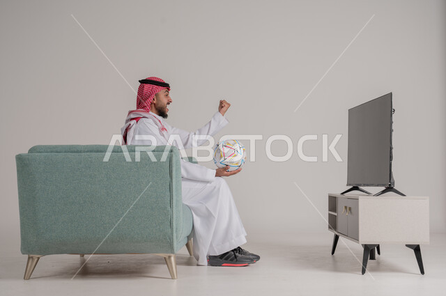  رفع اليد بإيماءات الحماس والسعادة لإحراز هدف، متابعة مباراة كرة القدم بتركيز وتمعن، بورتريه من الجانب لرجل عربي خليجي سعودي يجلس على كنبة مريحة امام التلفاز يمسك بالكرة ويشاهد لعب فريقه المفضل،تنمية شغف الاهتمامات الرياضية، الجسم بالطول الكامل، خلفية رمادية
