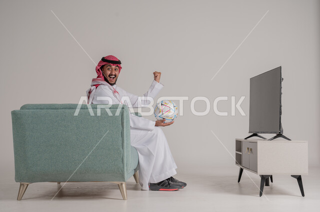 رفع اليد بإيماءات الحماس والسعادة لإحراز هدف، متابعة مباراة كرة القدم بتركيز وتمعن، بورتريه من الجانب لرجل عربي خليجي سعودي يجلس على كنبة مريحة امام التلفاز يمسك بالكرة ويشاهد لعب فريقه المفضل،تنمية شغف الاهتمامات الرياضية، الجسم بالطول الكامل، خلفية رمادية