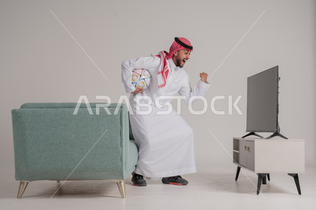 القفز فرحا وابتهاجا بإحراز هدف الفوز في الدوري، متابعة مباراة كرة القدم بتركيز وتمعن، بورتريه من الجانب لرجل عربي خليجي سعودي يستمتع بمشاهدة لعب فريقه المفضل، التفاعل مع الأهداف بسعادة وسرور، تنمية شغف الاهتمامات الرياضية، الجسم بالطول الكامل، خلفية رمادية