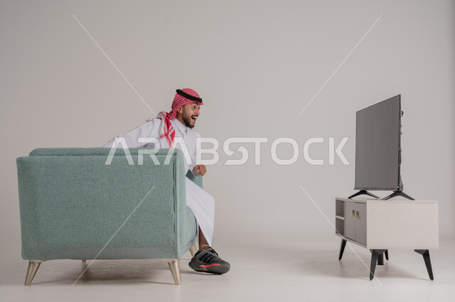 Gestures of enthusiasm, happiness and joy at scoring the winning goal, watching a football match with concentration and contemplation, side portrait of a Saudi Arabian Gulf man sitting on a comfortable sofa in front of the TV watching his favorite team play, developing passion for sports interests, full body, gray background
