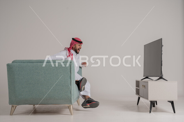 إيماءات الحماس والسعادة والفرح بإحراز هدف الفوز، متابعة مباراة كرة القدم بتركيز وتمعن، بورتريه من الجانب لرجل عربي خليجي سعودي يجلس على كنبة مريحة امام التلفاز ويشاهد لعب فريقه المفضل، تنمية شغف الاهتمامات الرياضية، الجسم بالطول الكامل، خلفية رمادية