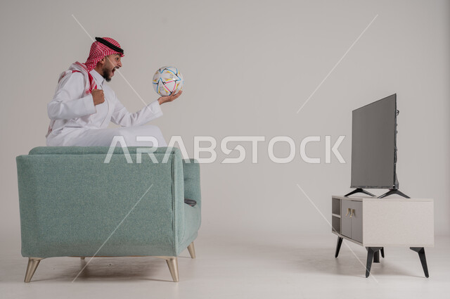 متابعة مباراة كرة القدم بتركيز وتمعن، بورتريه من الجانب لرجل عربي خليجي سعودي يجلس على كنبة مريحة امام التلفاز يمسك بالكرة ويشاهد لعب فريقه المفضل، رفع اليد بإيماءات الحماس والسعادة لإحراز هدف، تنمية شغف الاهتمامات الرياضية، الجسم بالطول الكامل، خلفية رمادية
